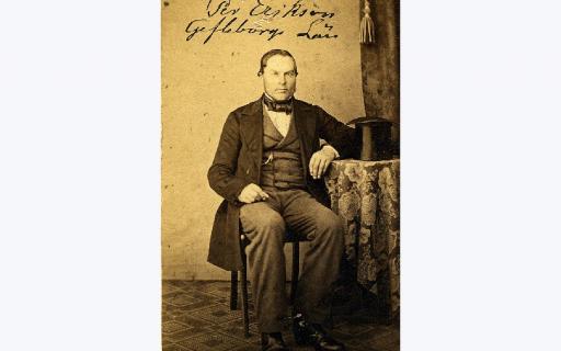 Bonderiksdagsmannen Pehr Ericsson (1815-92) i byxor väst och rock sitter med armen vilande mot runt bord där den höga hatten är placerad.