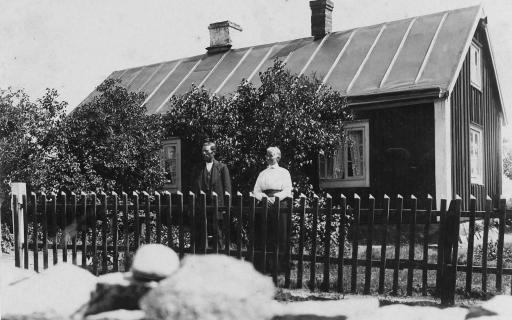 Gustav och Berta vid staketet framför sitt hus där stora syrenbuskar skymmer fönstren.