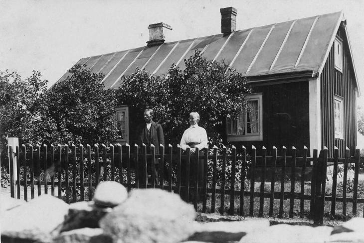 Gustav och Berta vid staketet framför sitt hus där stora syrenbuskar skymmer fönstren.